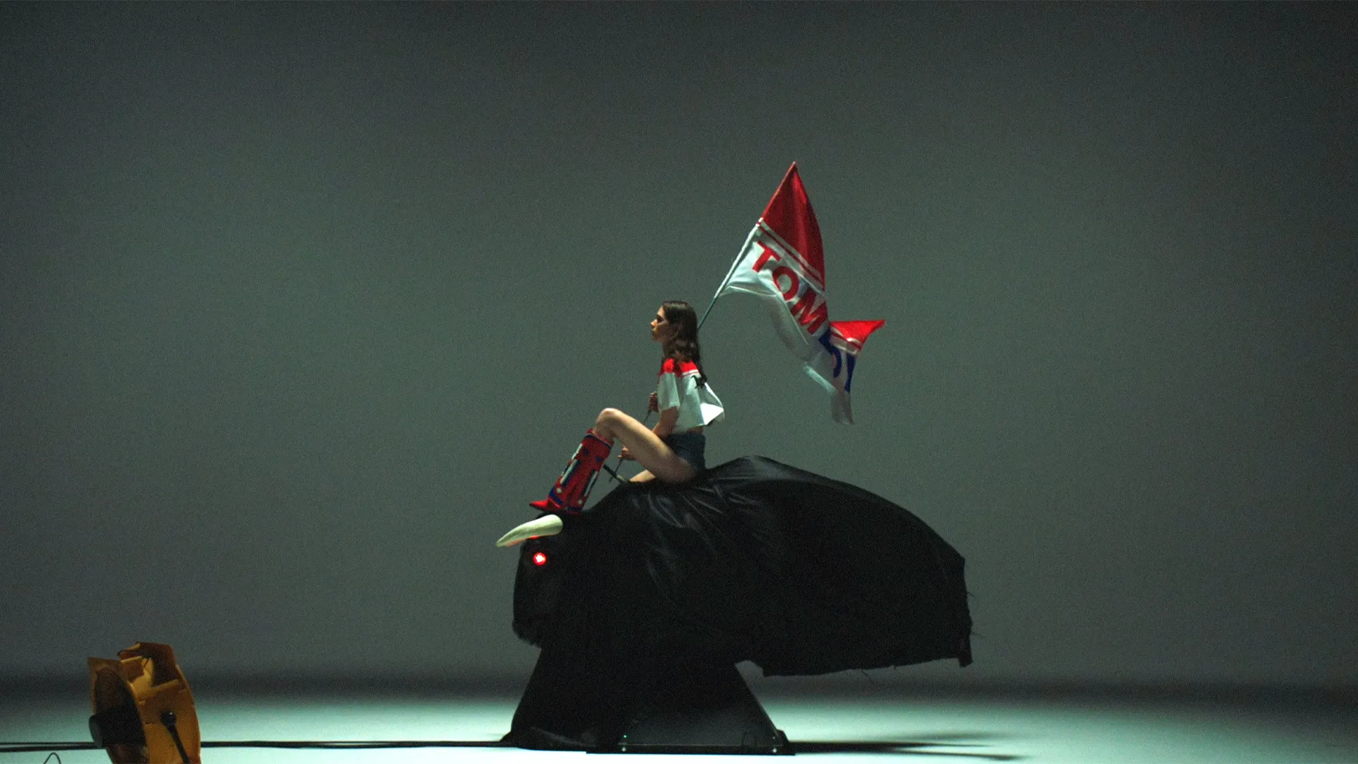 A young woman dressed in Tomboy clothing holding a flag is sitting on a mechanical bull.